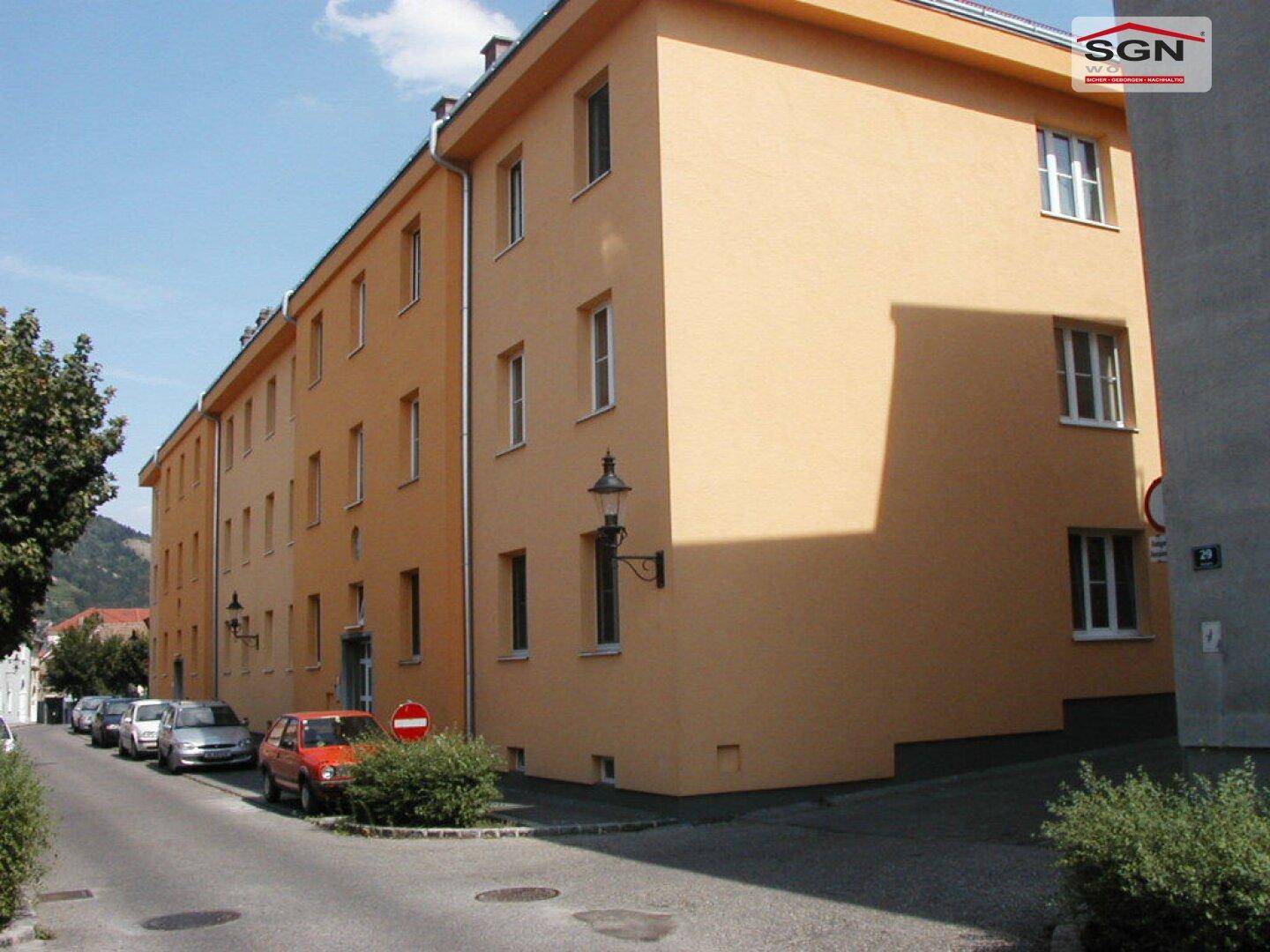 Ansicht eines Mehrfamilienhauses an einer Straße mit parkenden Autos und Bäumen.