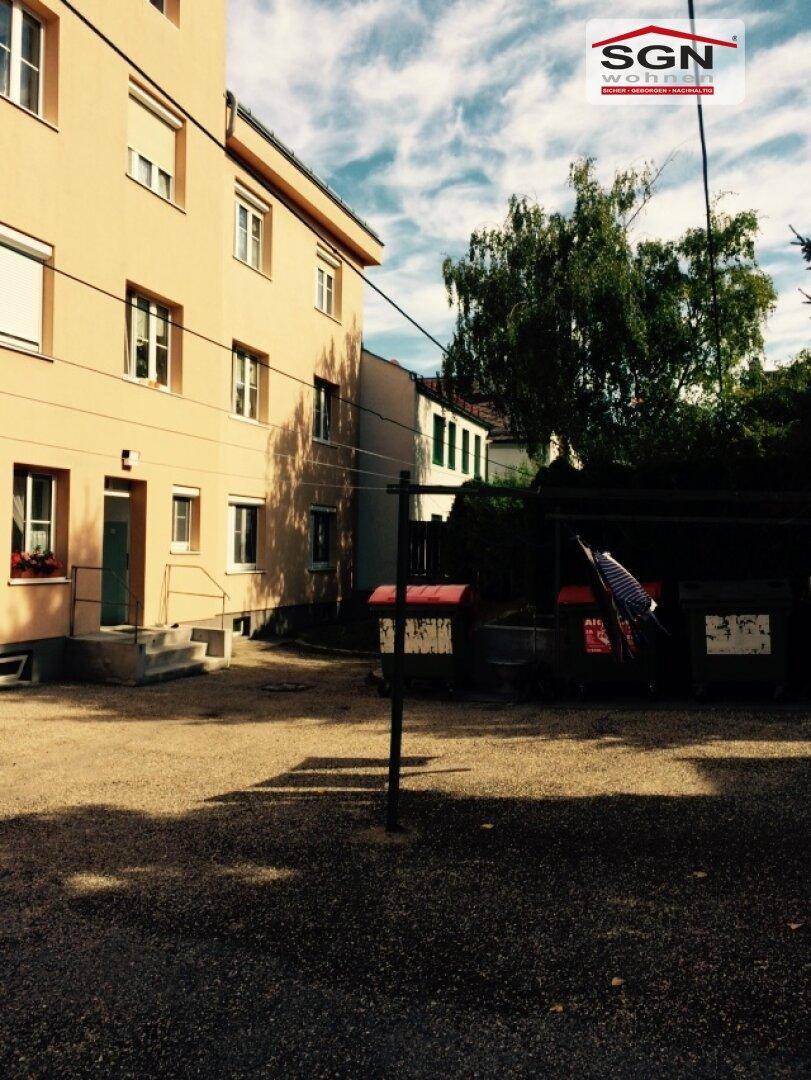 Rückansicht des Gebäudes mit einem Schotterplatz und Müllcontainern unter bewölktem Himmel.