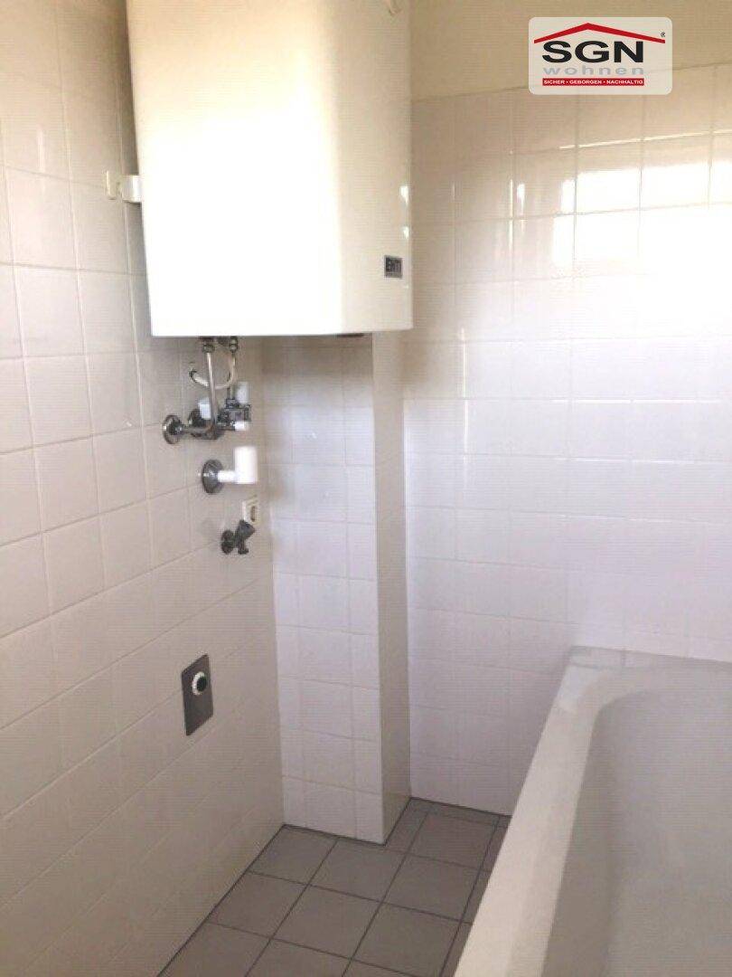 Badezimmer mit Warmwasserboiler und Badewanne, gefliest mit weißen Wand- und grauen Bodenfliesen.