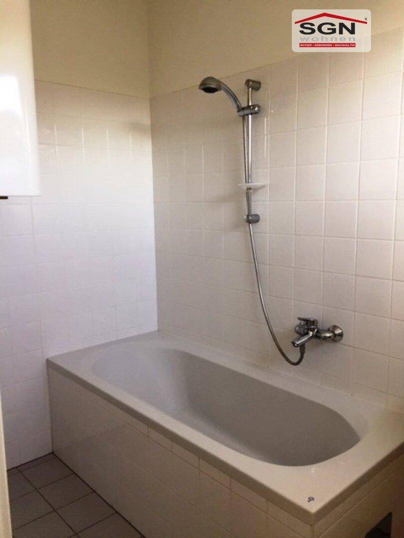 Badezimmer mit Badewanne und Duschvorrichtung, komplett gefliest in Weiß und Grau, funktional und sauber.