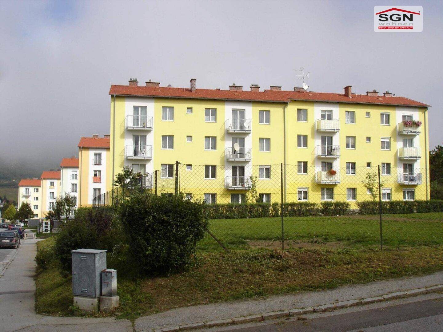 Mehrfamilienhaus mit gelber Fassade und mehreren Balkonen in einer gepflegten Wohnsiedlung mit Grünflächen.