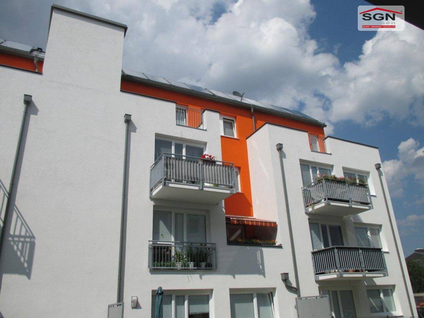 Detailansicht der modernen Fassade mit orangefarbenen Elementen, Balkonen und Solarpaneelen auf dem Dach.