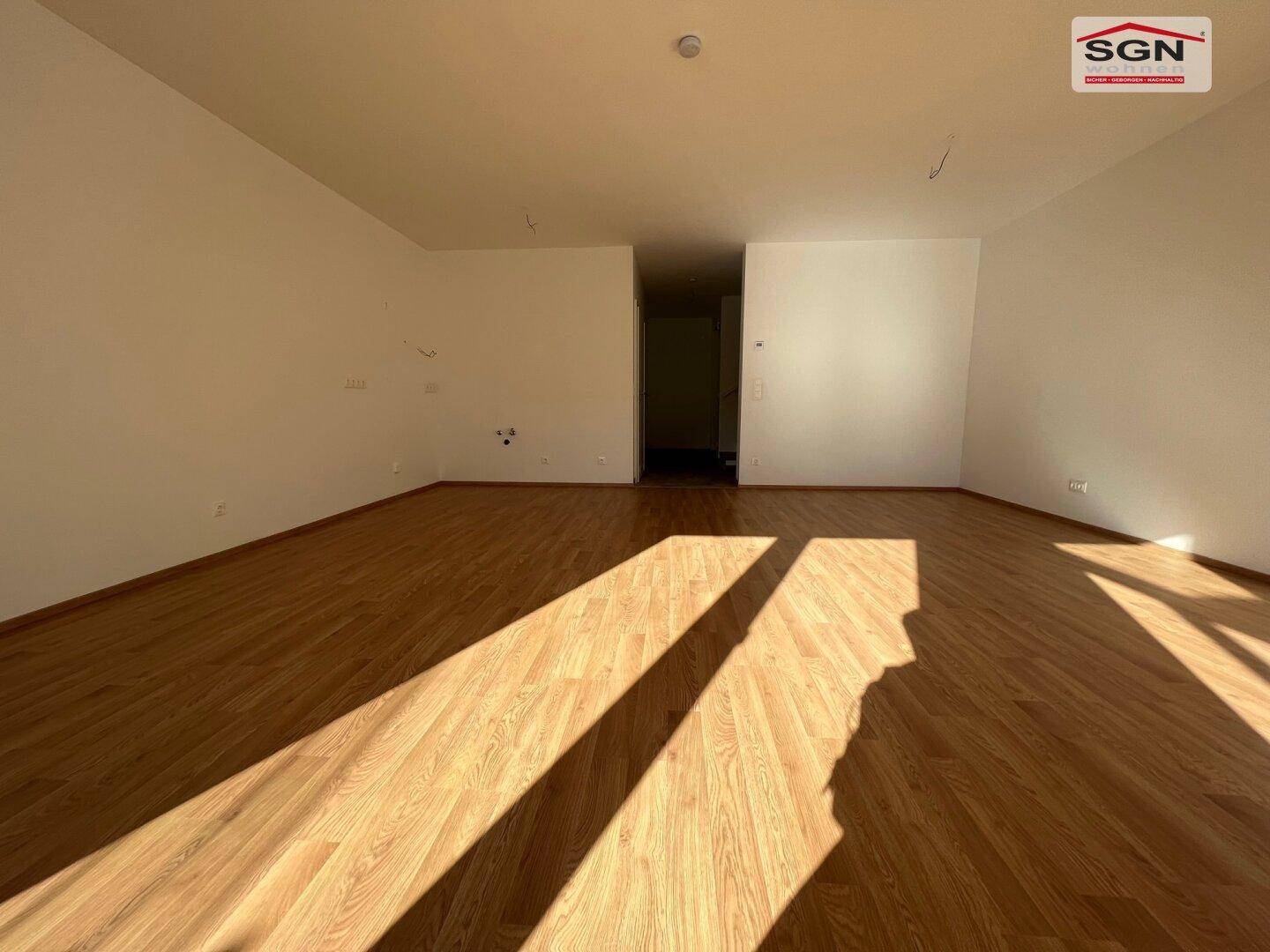 Geräumiges Wohnzimmer mit hellem Laminatboden und viel natürlichem Licht durch große Fenster.
