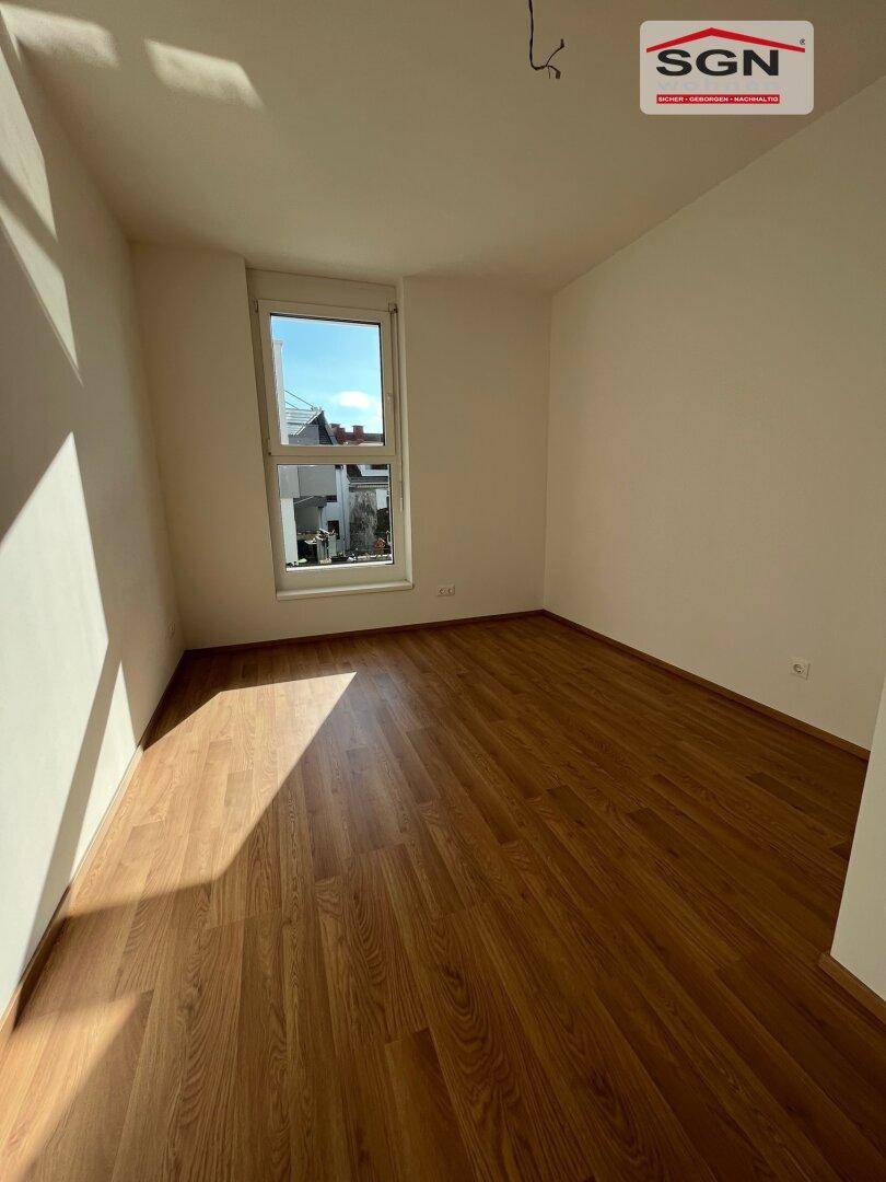 Helles, leeres Zimmer mit Laminatboden und einem Fenster, das viel Tageslicht hereinlässt.