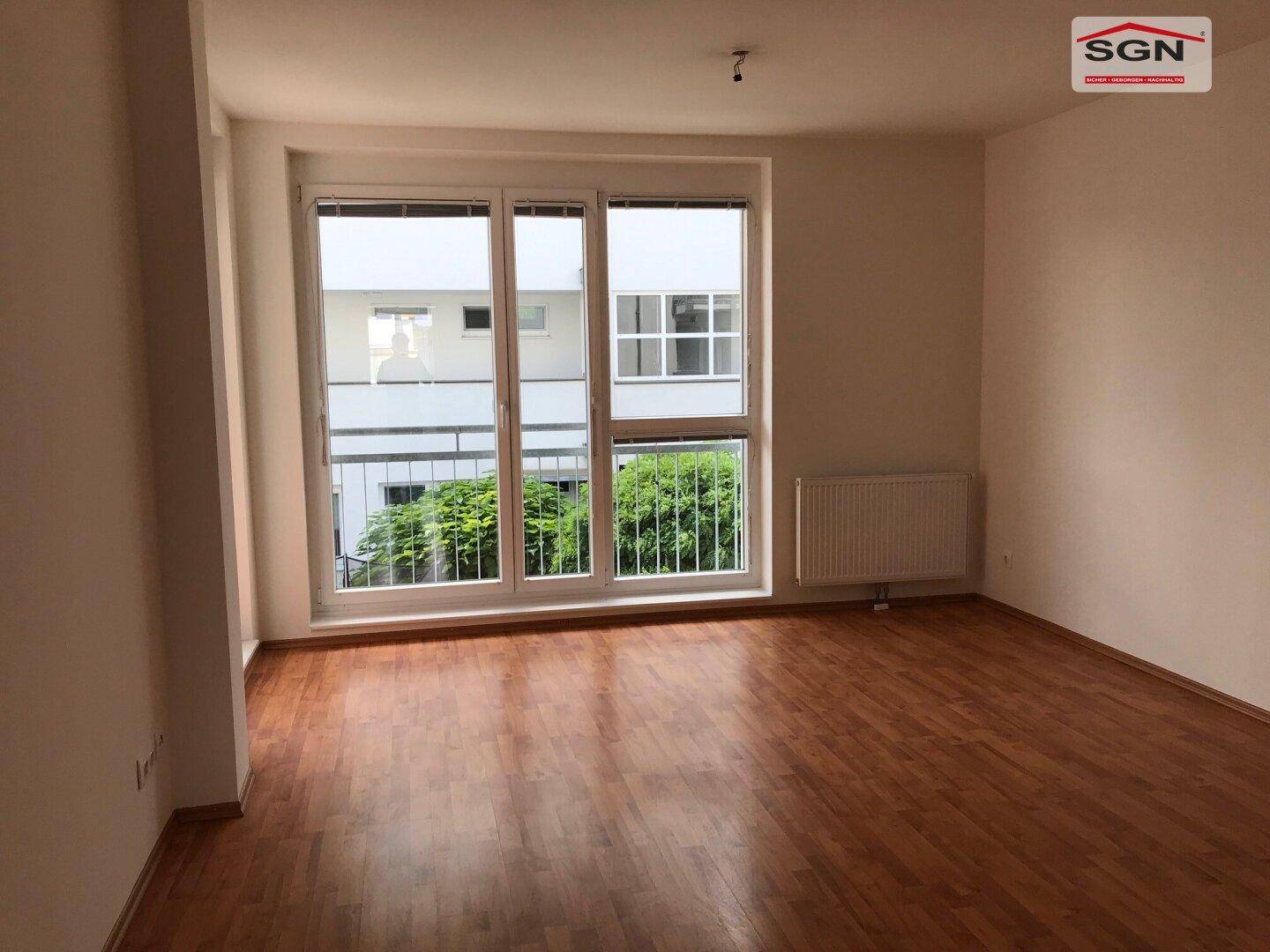 Helles Wohnzimmer mit Laminatboden und großem Fenster, das viel Tageslicht hereinlässt.