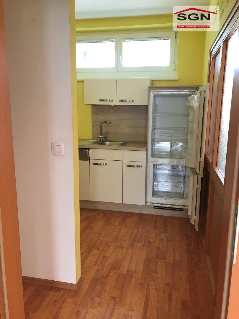 Kleine Einbauküche mit weißen Schränken, Spüle und Kühlschrank in einem hellgelben Raum.
