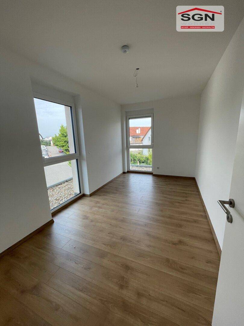 Leeres Zimmer mit Laminatboden und zwei großen Fenstern, die viel Tageslicht hereinlassen.
