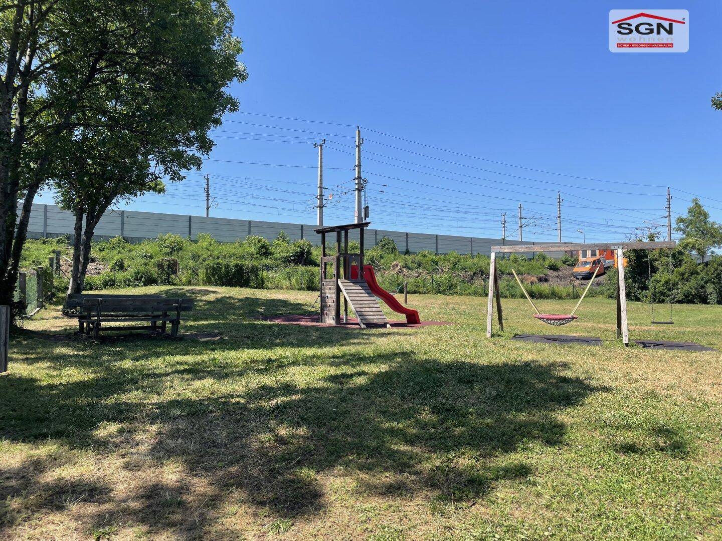 Kinderspielplatz mit Rutsche und Schaukeln, umgeben von Grünflächen und Bäumen.