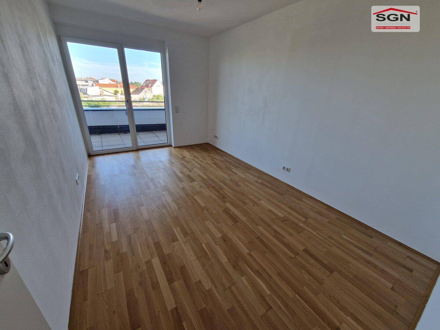 Leeres Zimmer mit Parkettboden und Zugang zu einem Balkon, bietet viel Tageslicht.