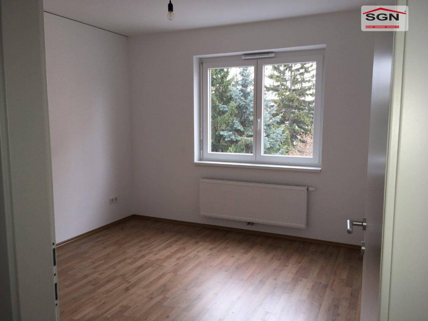 Unmöbliertes Schlafzimmer mit Holzboden, Fenster und Blick auf Bäume.