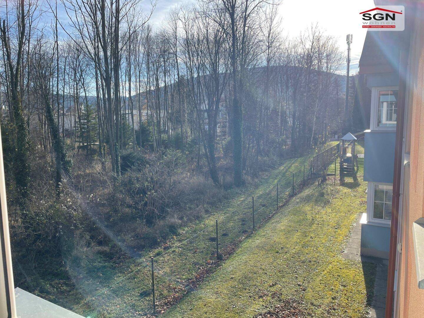 Blick aus dem Fenster auf einen Spielplatz und eine bewaldete Böschung mit vielen Bäumen.