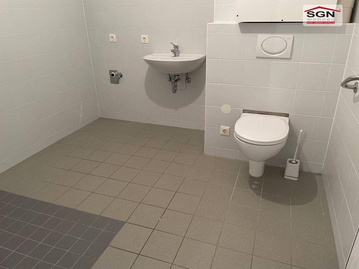Helles, gefliestes WC mit wandmontierter Toilette, Waschbecken und Haltegriffen für Barrierefreiheit.