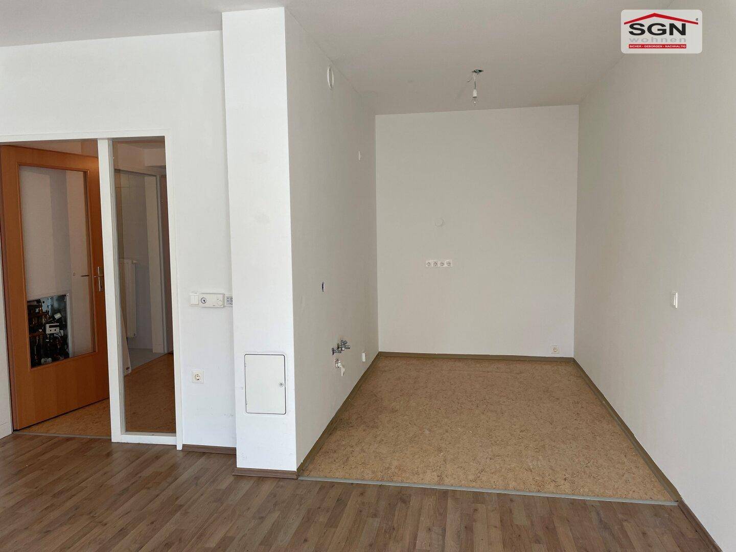 Geräumiger, leerer Wohnbereich mit hellem Holzboden und vorbereiteten Anschlüssen für eine Küche.