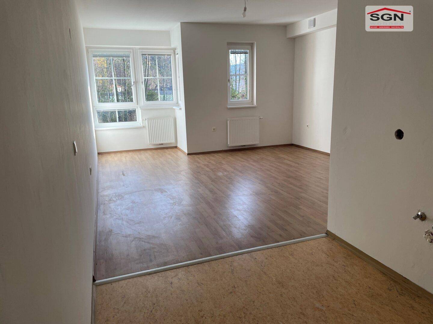 Leerer Wohnraum mit großen Fenstern, die viel Tageslicht hereinlassen, und Holzboden.