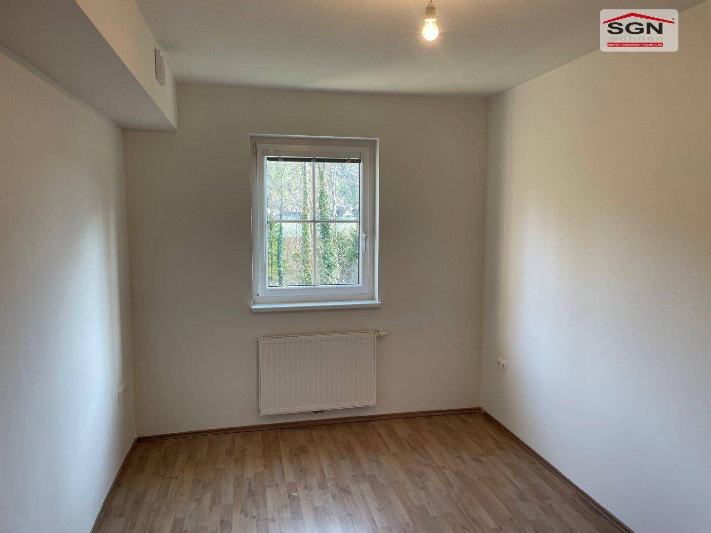 Helles, leeres Schlafzimmer mit Holzboden und einem Fenster mit Blick ins Grüne.