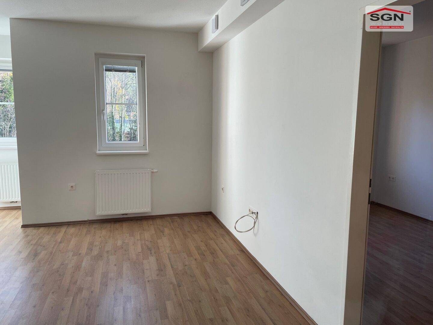 Heller, leerer Wohnraum mit Holzboden und einem Fenster, das Tageslicht hereinlässt.