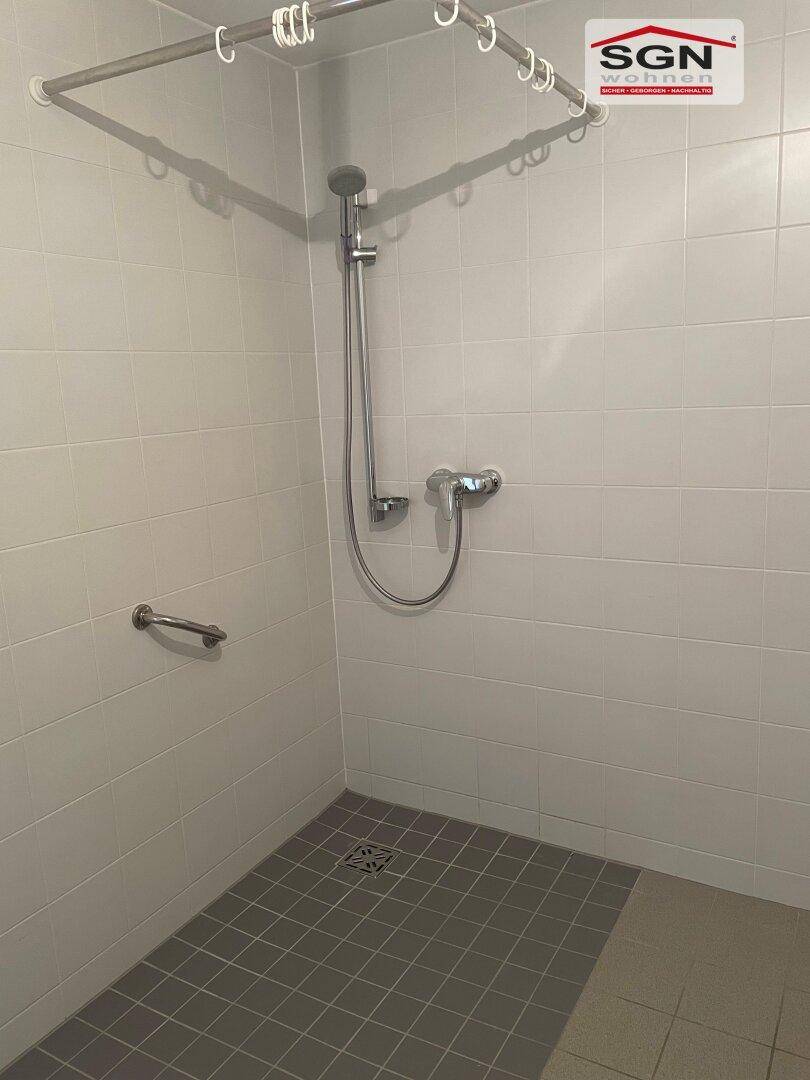 Barrierefreie Dusche mit Haltegriff und Duschvorhangstange, komplett gefliest in hellen Farben.