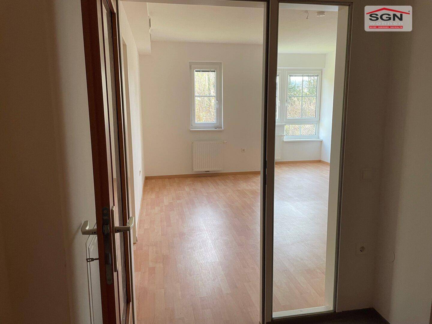 Blick durch eine Glastür in einen hellen, leeren Raum mit Laminatboden und großen Fenstern.