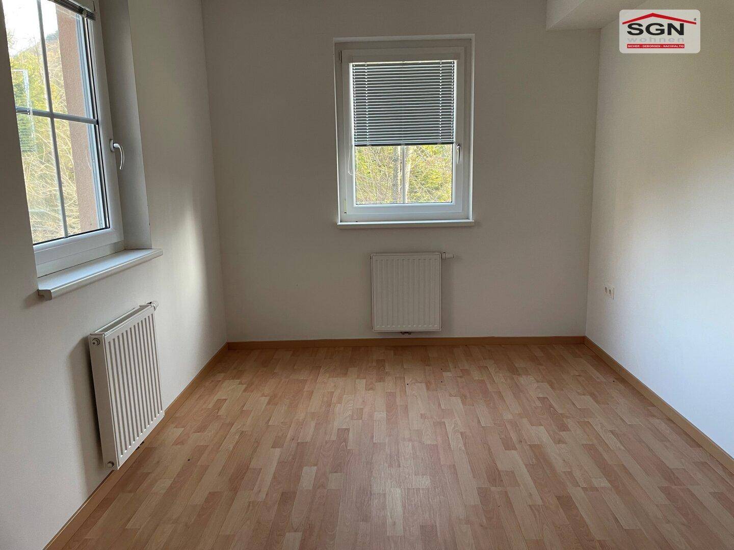 Leeres Schlafzimmer mit Laminatboden und einem Fenster mit Jalousien, das viel Tageslicht hereinlässt.