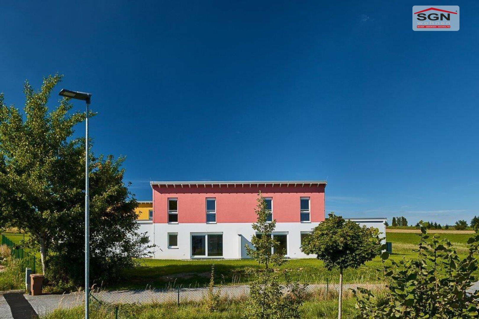Vorderansicht eines modernen Neubaus mit roter Fassade, großen Fenstern und grünem Vorgarten.