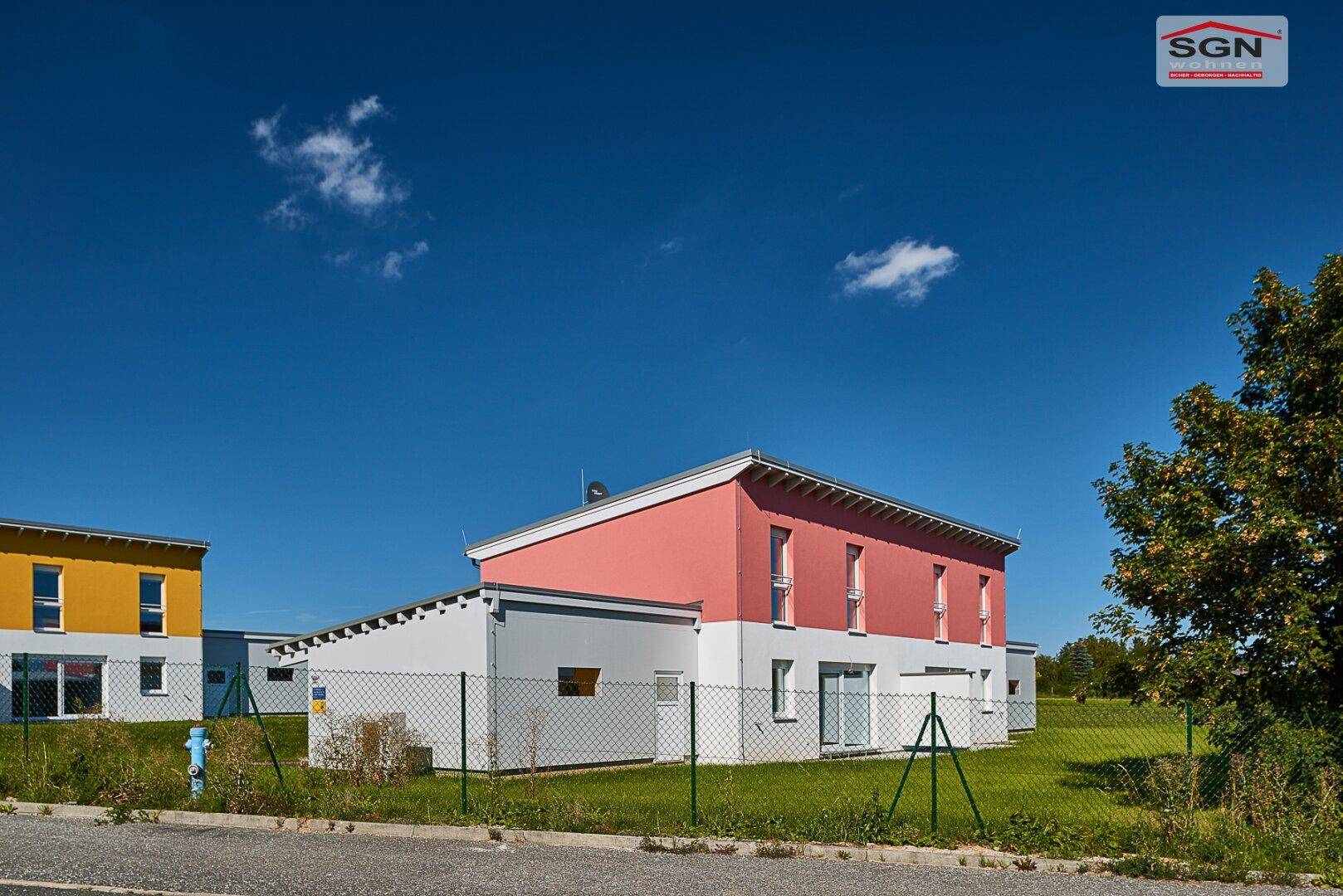 Seitenansicht des Neubaus mit roter Fassade und angrenzendem Gartenbereich unter blauem Himmel.