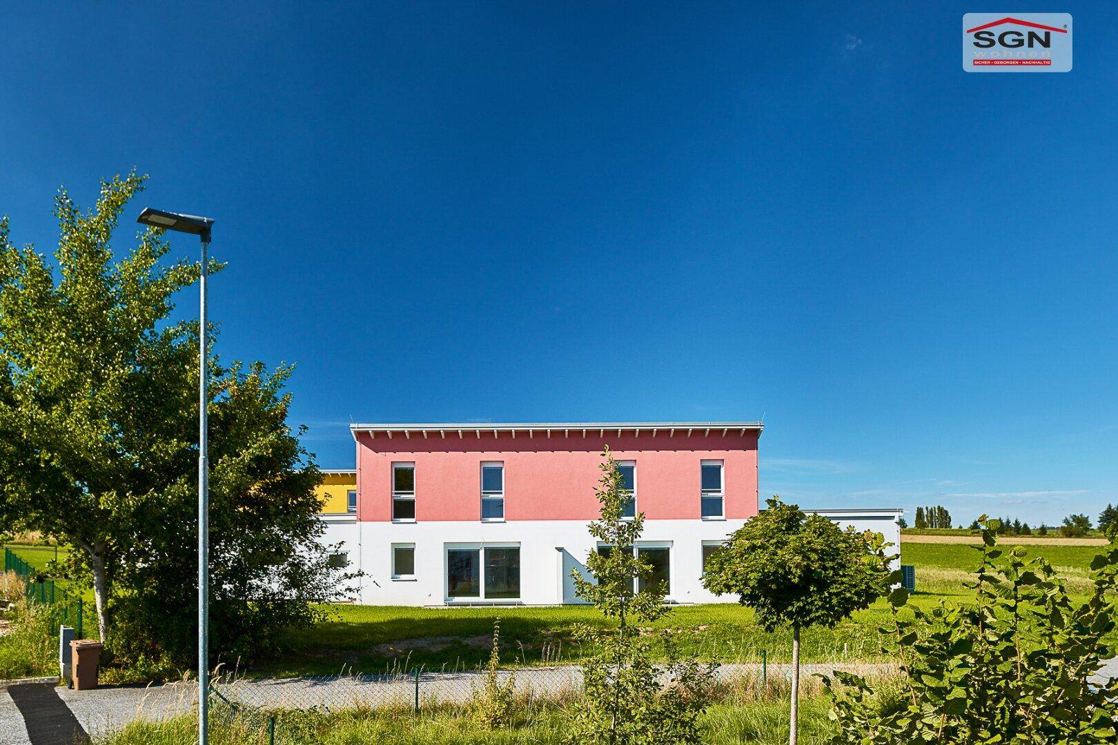 Vorderansicht eines modernen Neubaus mit roter Fassade, großen Fenstern und grünem Vorgarten.