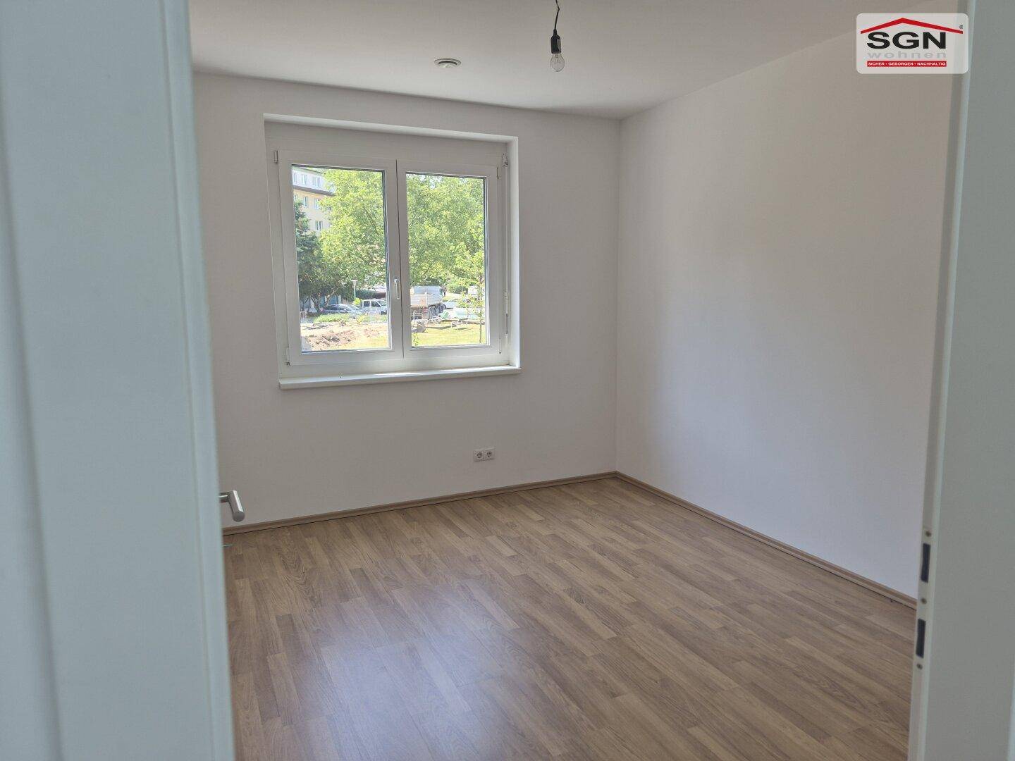 Helles, leeres Schlafzimmer mit Laminatboden und großem Fenster mit Blick ins Grüne.