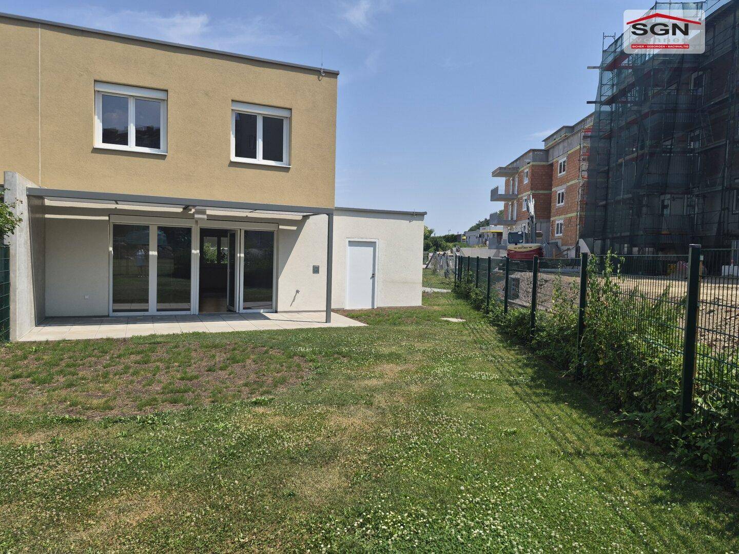 Modernes Reihenhaus mit Garten und angrenzender Baustelle unter blauem Himmel.
