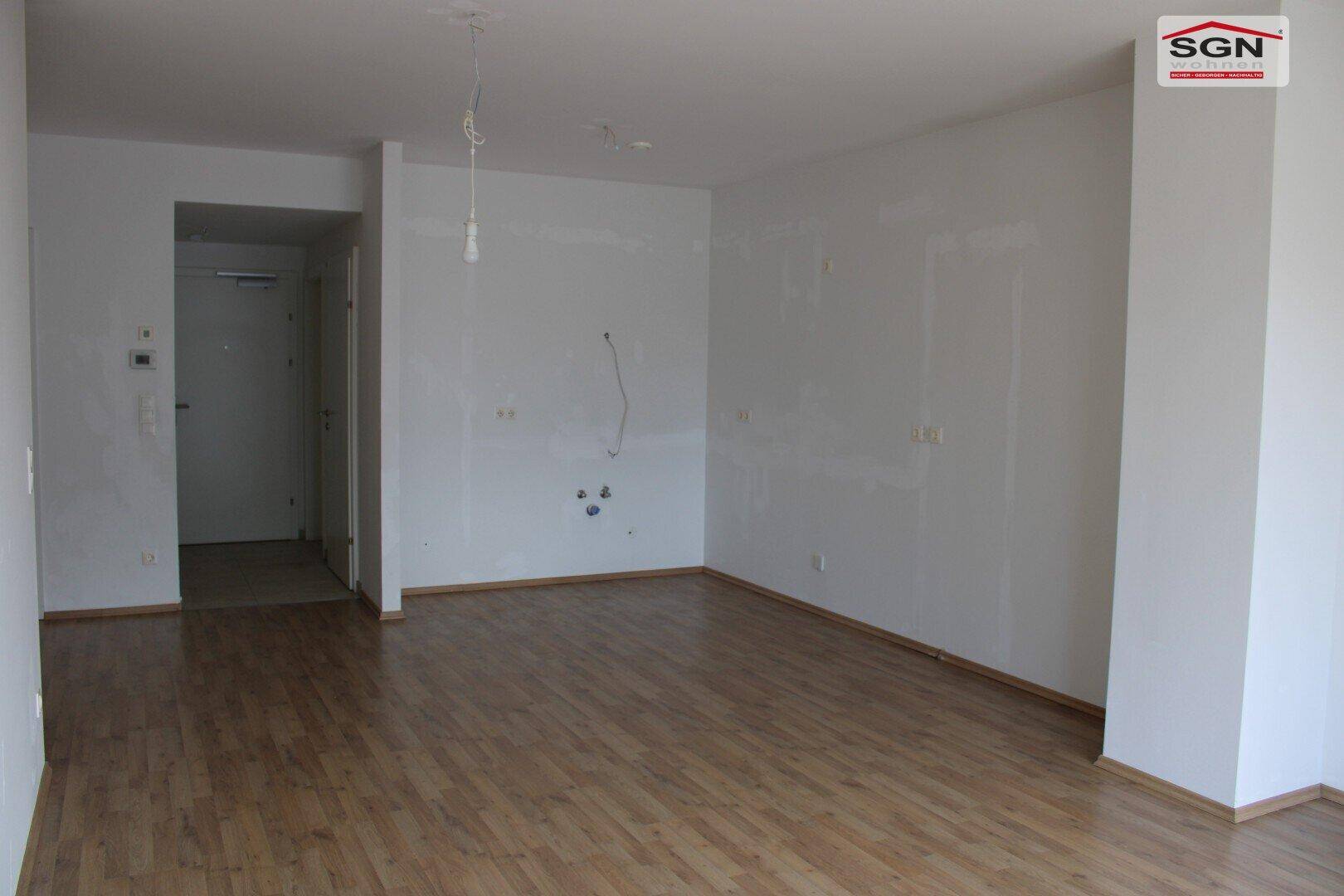 Geräumiger, heller Wohnbereich mit Holzboden und Zugang zu einem Flur, unmöbliert.