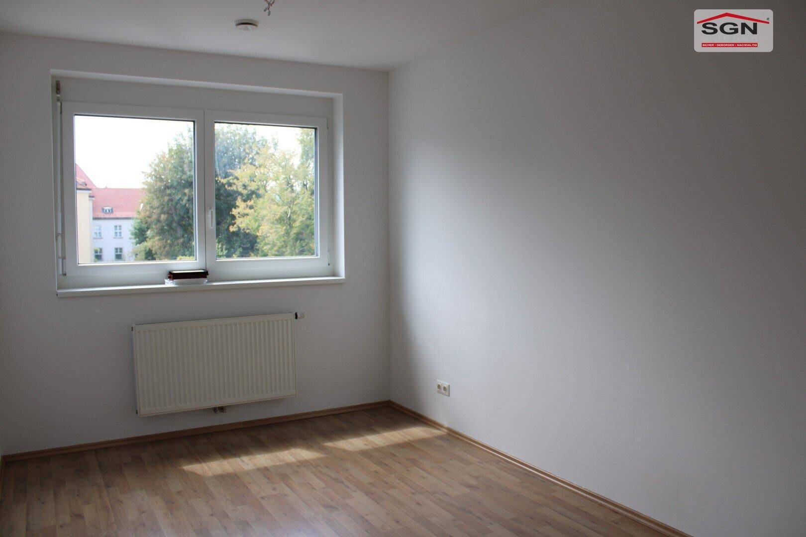Helles Schlafzimmer mit Holzboden, großem Fenster und Heizkörper, bietet viel Tageslicht.