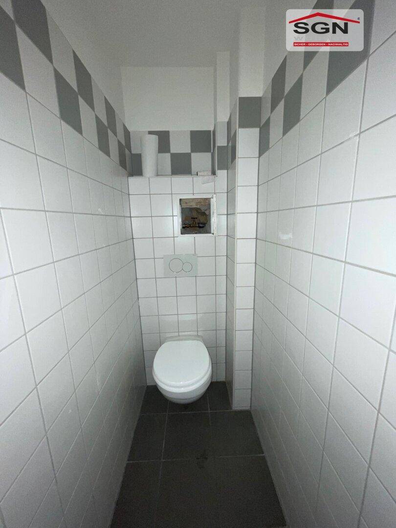Schmale Toilette mit weißen Fliesen und einem grauen Schachbrettmuster an der Wand, funktional und sauber.