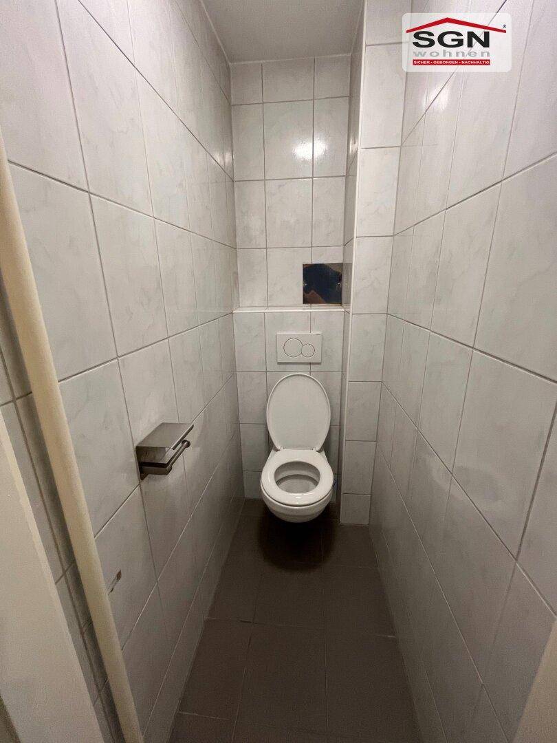 Schmale, geflieste Toilette mit Wand-WC und dunklem Boden, funktional und sauber.