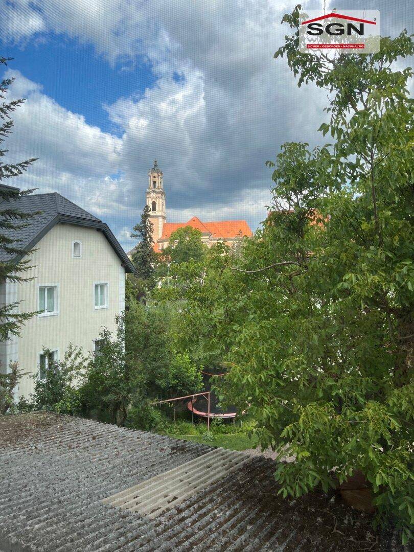 Blick aus dem Fenster auf grüne Bäume und eine Kirche mit rotem Dach in der Ferne.
