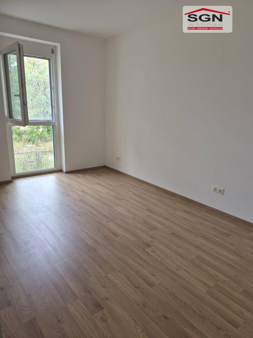 Leeres Zimmer mit Laminatboden und einem großen Fenster, das viel Tageslicht hereinlässt.