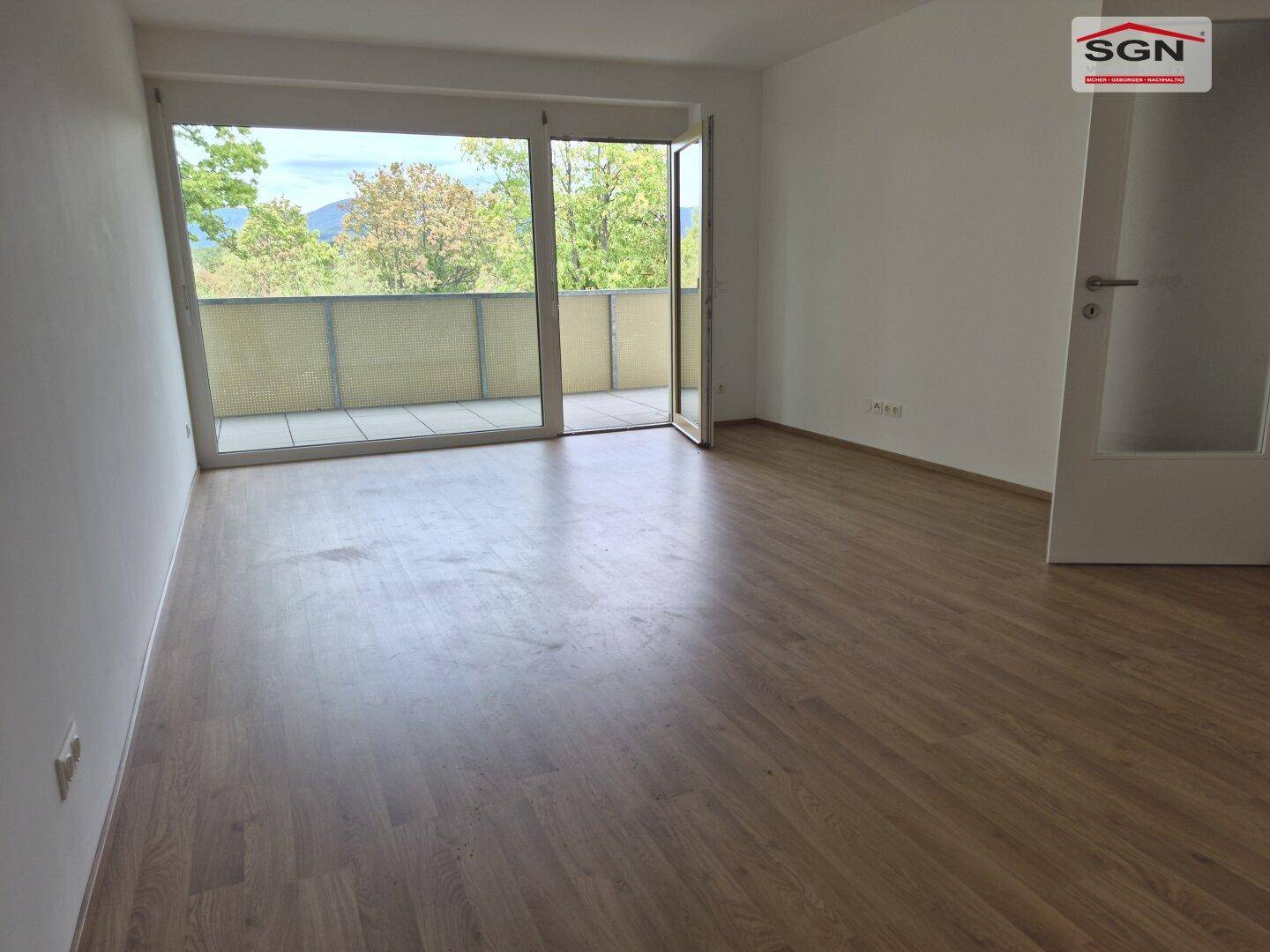 Helles Wohnzimmer mit Laminatboden und großem Balkonzugang, der einen weiten Blick ins Grüne bietet.