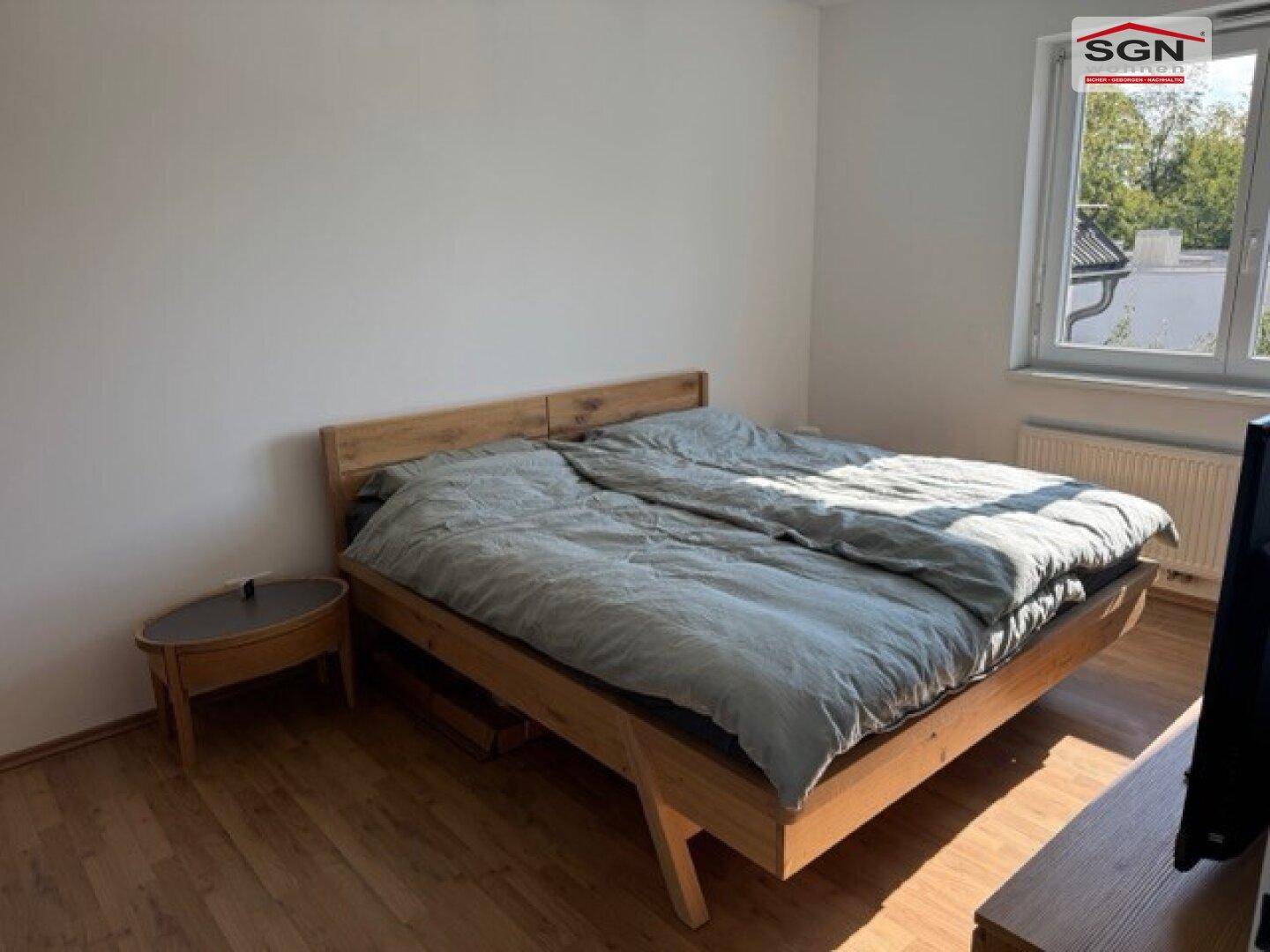 Helles Schlafzimmer mit Holzbett, Nachttisch und Fenster mit Blick nach draußen.
