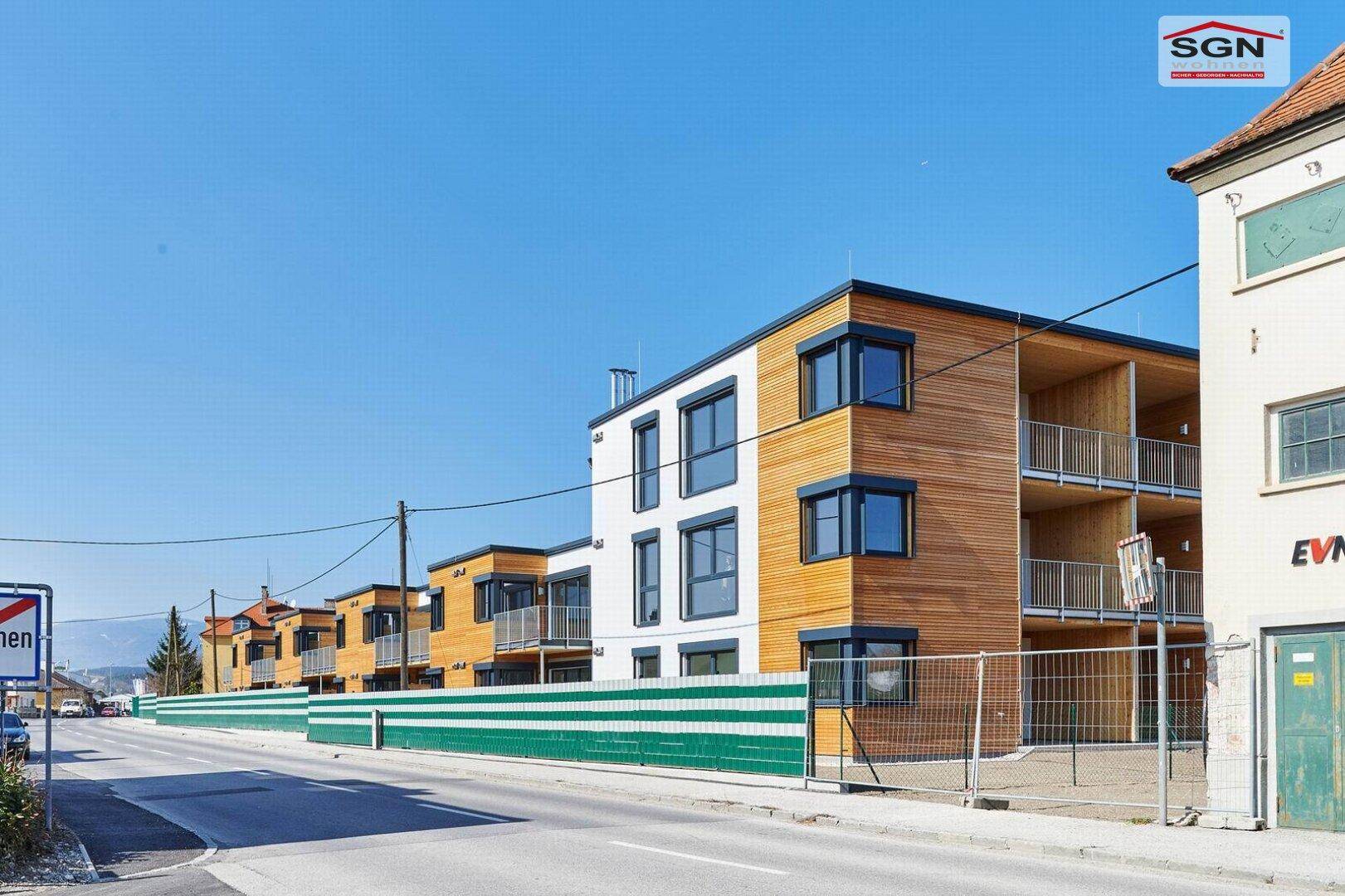 Moderne Wohngebäude mit Holzfassaden und Balkonen, von der Straße aus gesehen, mit einem grünen Zaun.