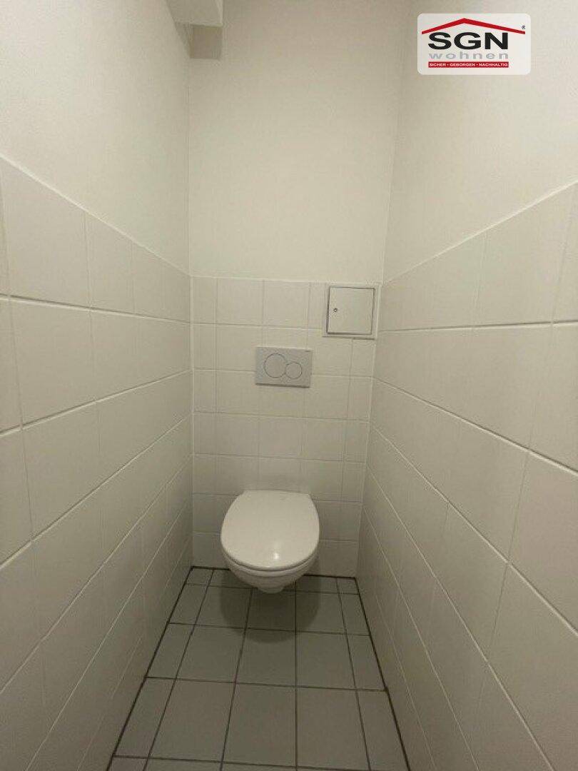 Separates WC mit wandhängender Toilette und weißen Wand- und Bodenfliesen.