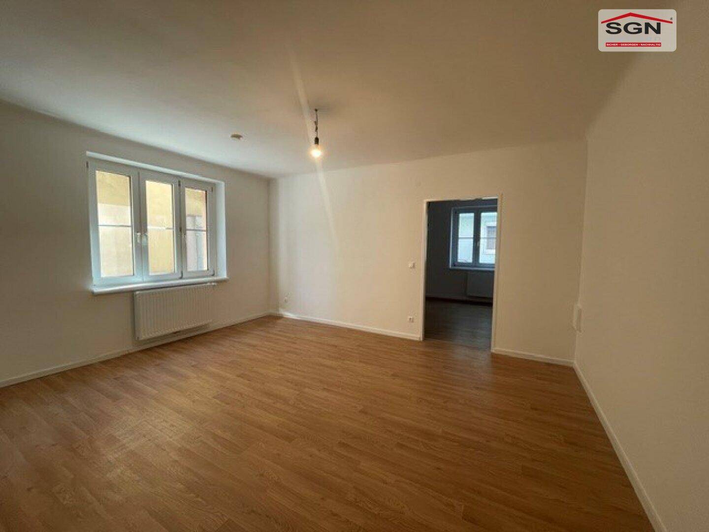 Geräumiges Wohnzimmer mit Holzfußboden, großen Fenstern und Zugang zu einem weiteren Raum.