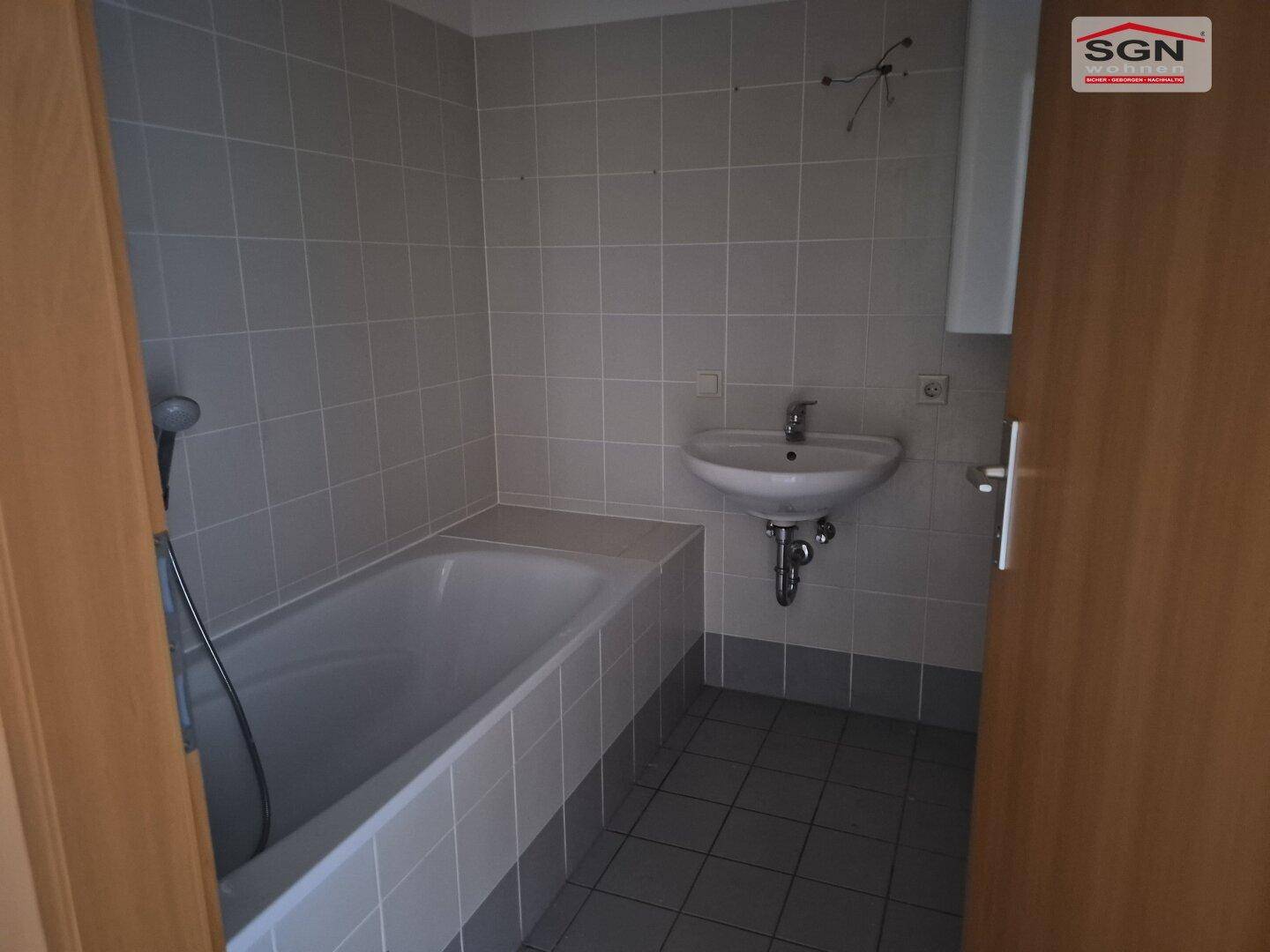 Badezimmer mit grauen Fliesen, einer Badewanne mit Duschkopf und einem einzelnen Waschbecken.