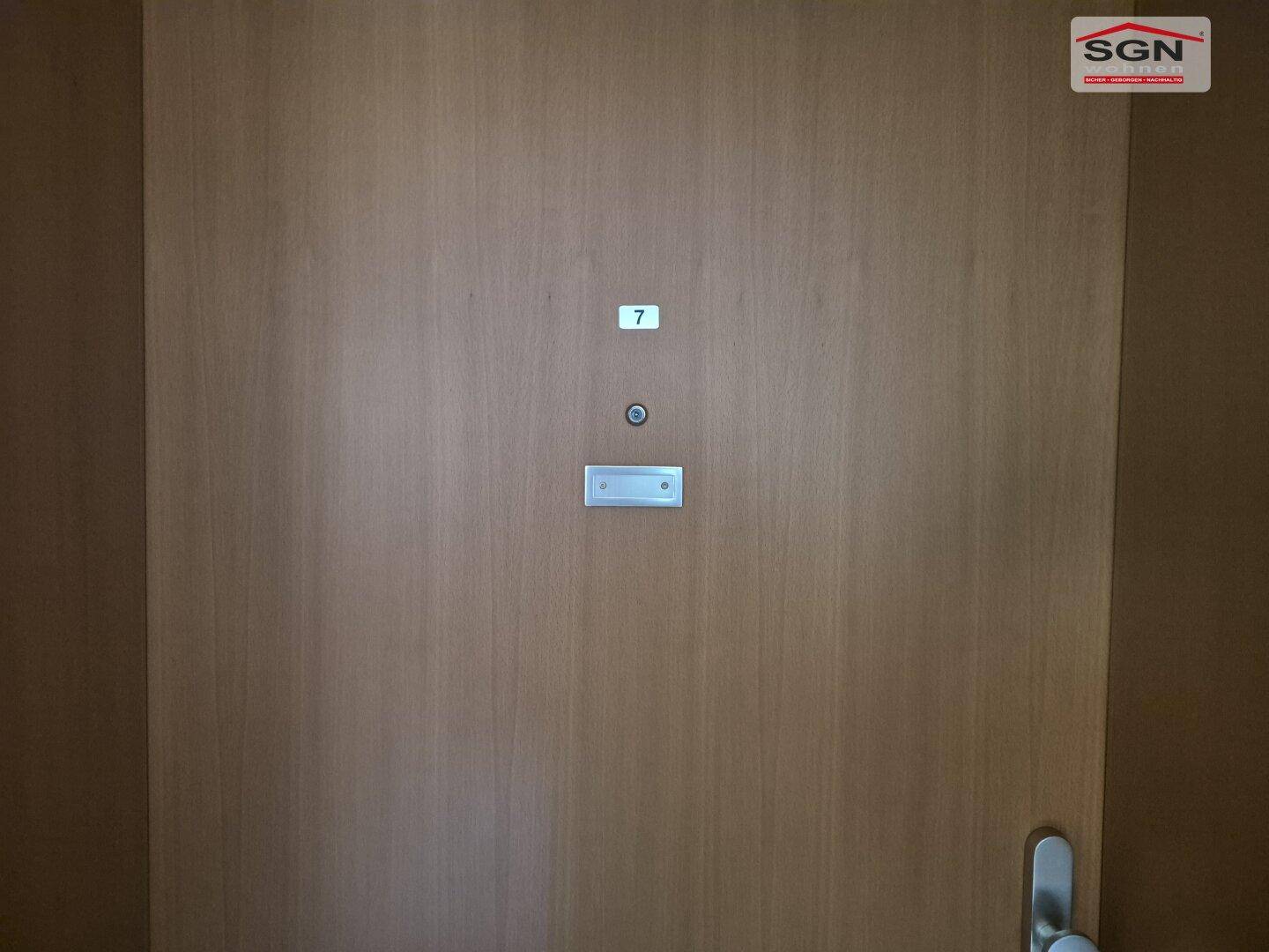 Eine schlichte Holztür mit der Nummer 7 und einem Türspion, wahrscheinlich der Eingang zu einer Wohnung.