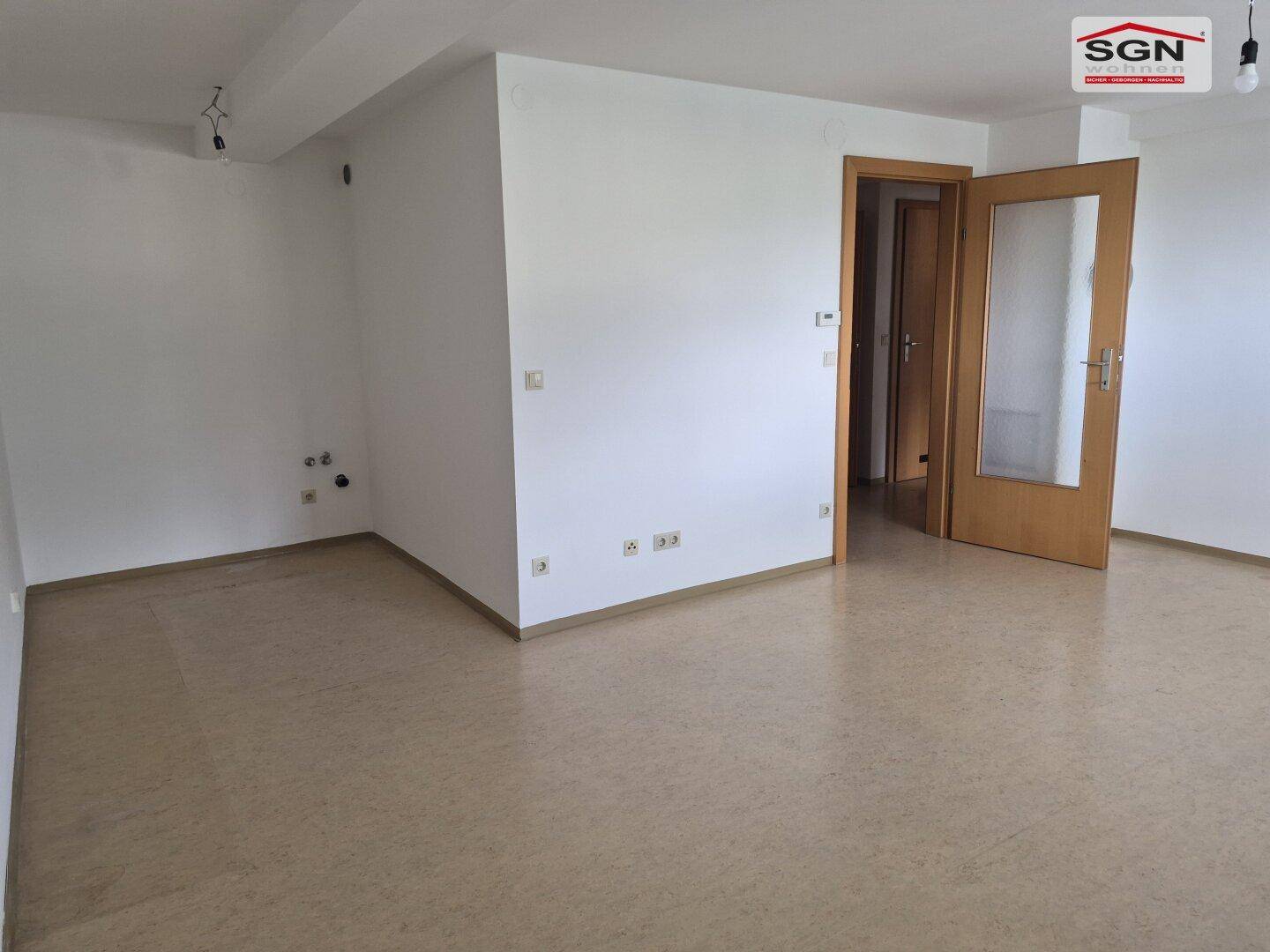 Leerer Wohnraum mit hellbraunem Boden, weißen Wänden und einer Holztür mit Glaseinsatz.