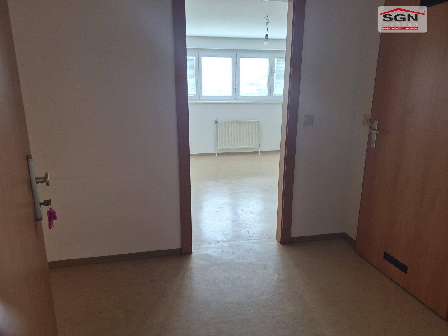 Blick aus einem Flur in einen hellen, leeren Raum mit Fenster und Heizkörper, der Boden ist hellbraun.