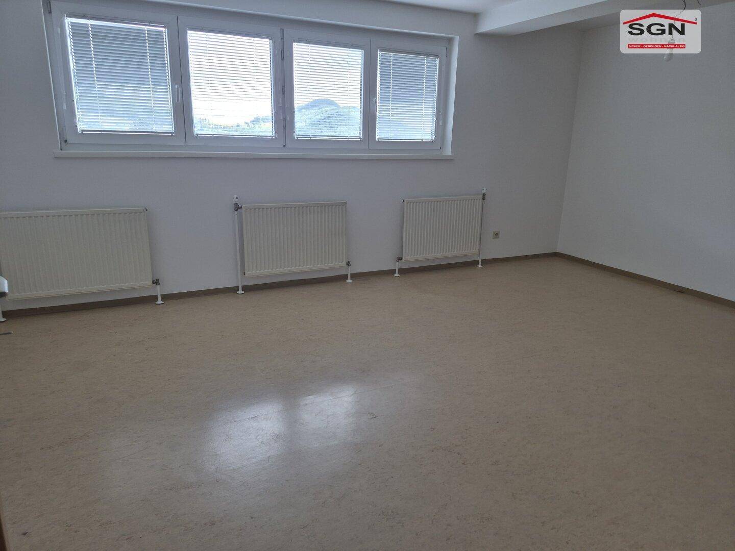Heller, leerer Raum mit hellbraunem Boden, weißen Wänden und einem großen Fenster mit Jalousien und Heizkörpern.