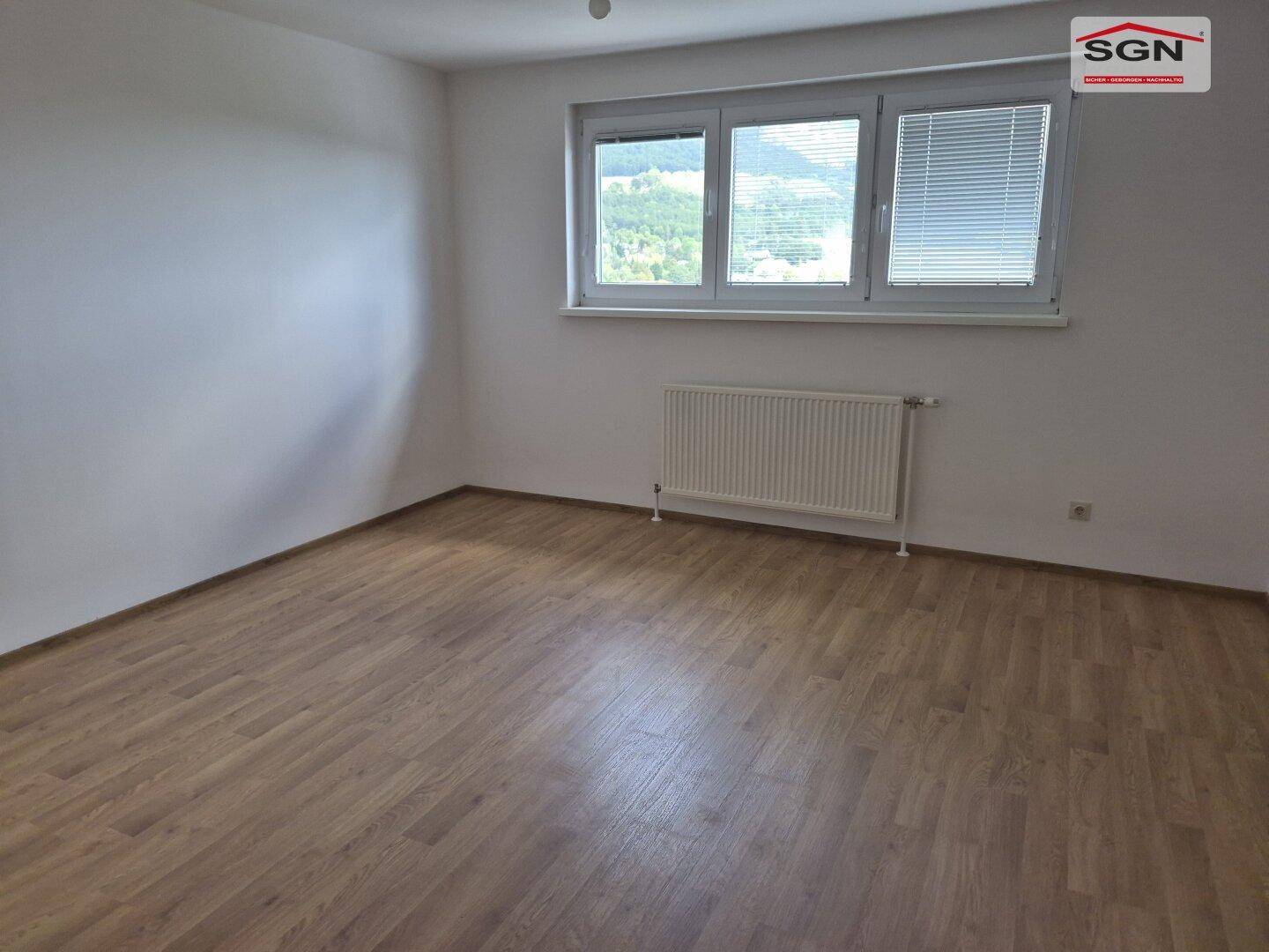Leeres Schlafzimmer mit Holzplankenboden, weißen Wänden und einem Fenster mit Jalousien und Heizkörper.