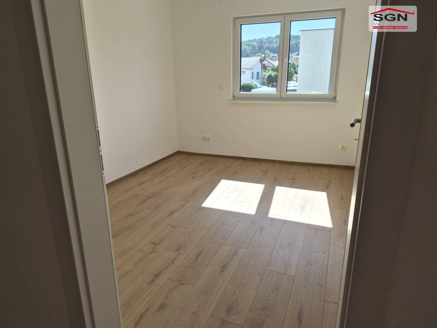 Leeres Schlafzimmer mit hellem Laminatboden und Fenster mit Blick ins Grüne.