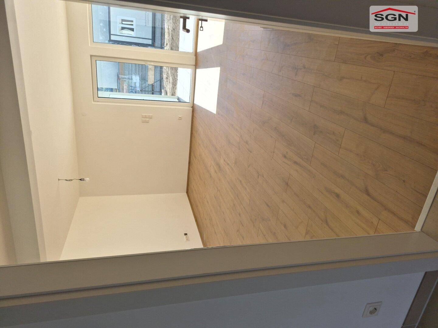 Heller, leerer Wohnraum mit Laminatboden und großen Fenstern, die viel Sonnenlicht hereinlassen.