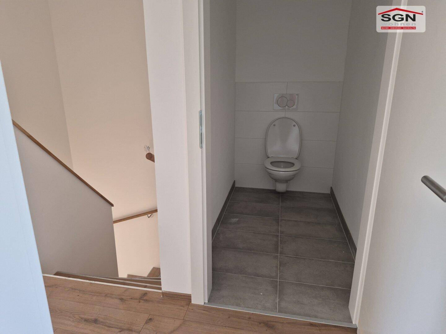 Kleines WC mit Wand-WC und grauen Fliesen, direkt neben dem Treppenaufgang gelegen.