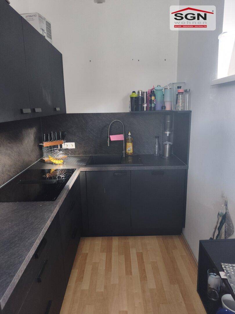 L-förmige Küche mit schwarzen Unterschränken, Spüle, Kochfeld und hellem Laminatboden.