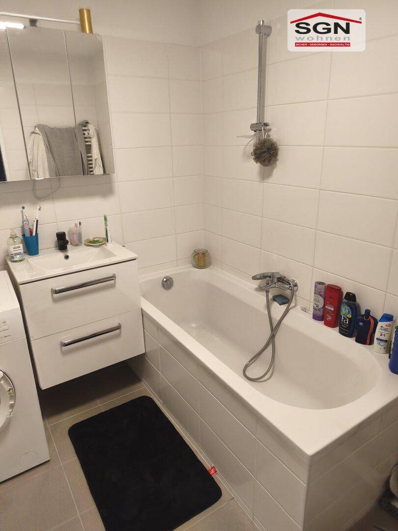Helles Badezimmer mit Badewanne, Waschtisch und weißen Fliesen an Wänden und Boden.